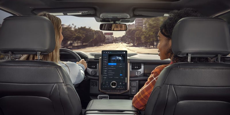 Two people in the front seats of a car, viewed from behind, with a large center touchscreen and road ahead.