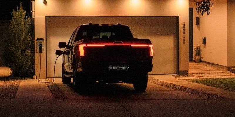 Dark electric pickup truck charging at home at night, with illuminated red taillights and light bar.