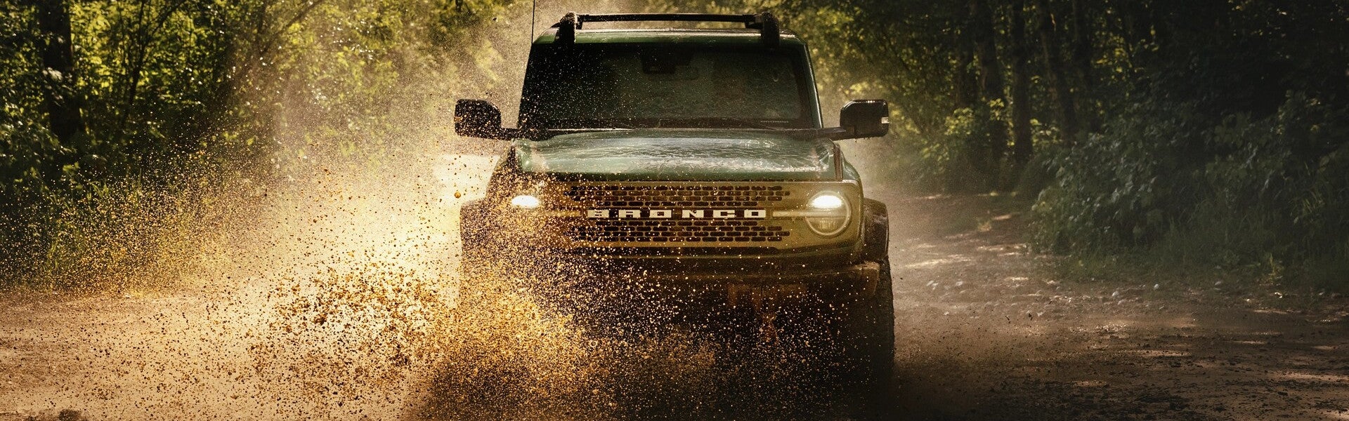 Dark green Ford Bronco driving on a muddy dirt road, splashing mud and water forward, headlights on, through a forest.