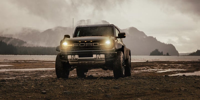 Dark Ford Bronco SUV with headlights on, parked on a muddy shore with a lake and misty mountains in the background.