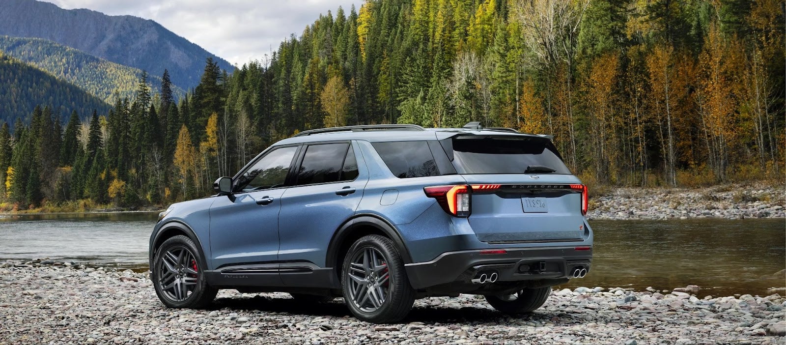 Blue SUV parked on a gravel riverbank with mountains and fall foliage in the background.