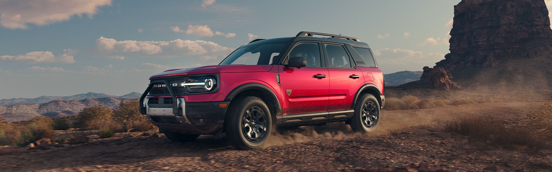 Red SUV driving on a dusty dirt road in a desert landscape with mountains and a large rock formation.