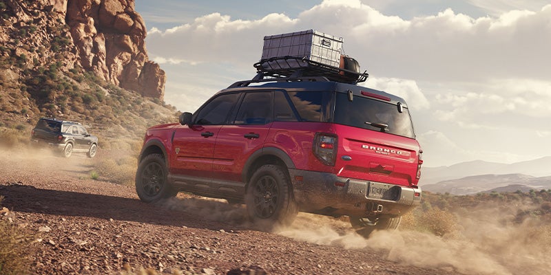 Two red and black SUVs kick up dust while off-roading on a dirt trail in a rocky, mountainous desert.