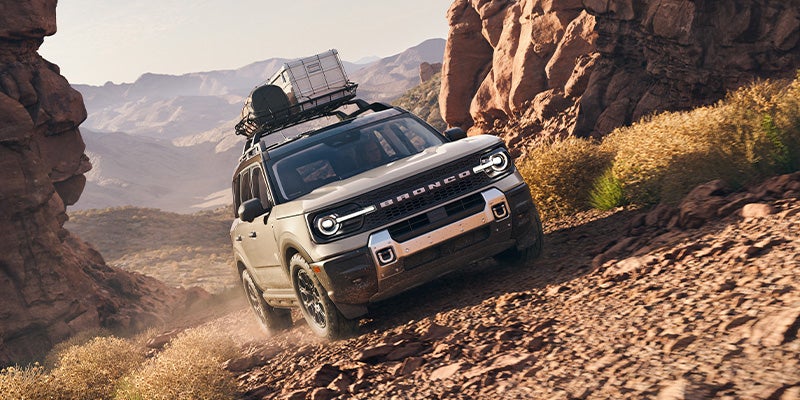 Light gray Ford Bronco Sport SUV with roof cargo driving down a rocky desert path surrounded by mountains."