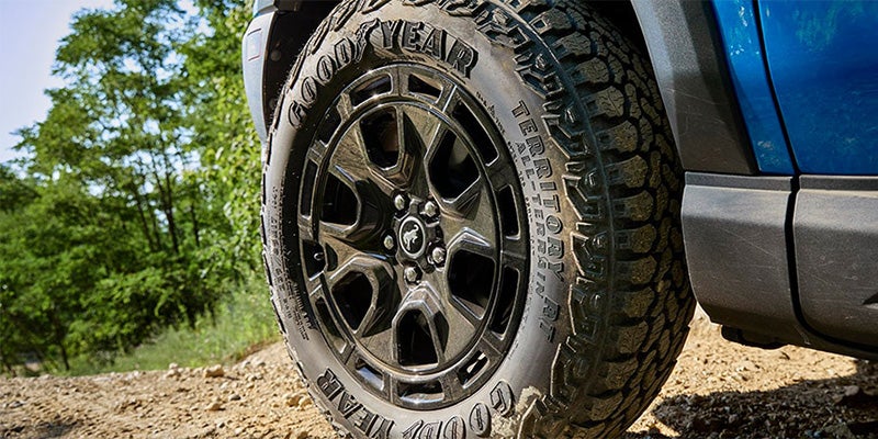 Close-up of a vehicle's Goodyear Territory All-Terrain tire and dark wheel with a Bronco logo, next to a blue car body.