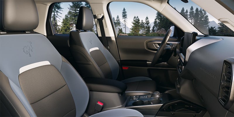 Car interior with gray and black two-tone seats featuring a Bronco logo, dashboard, and view of mountains outside.