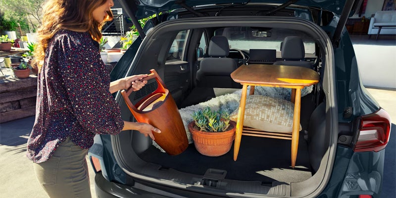 Woman loading a small wooden table, potted plant, and other items into the open trunk of a dark SUV.