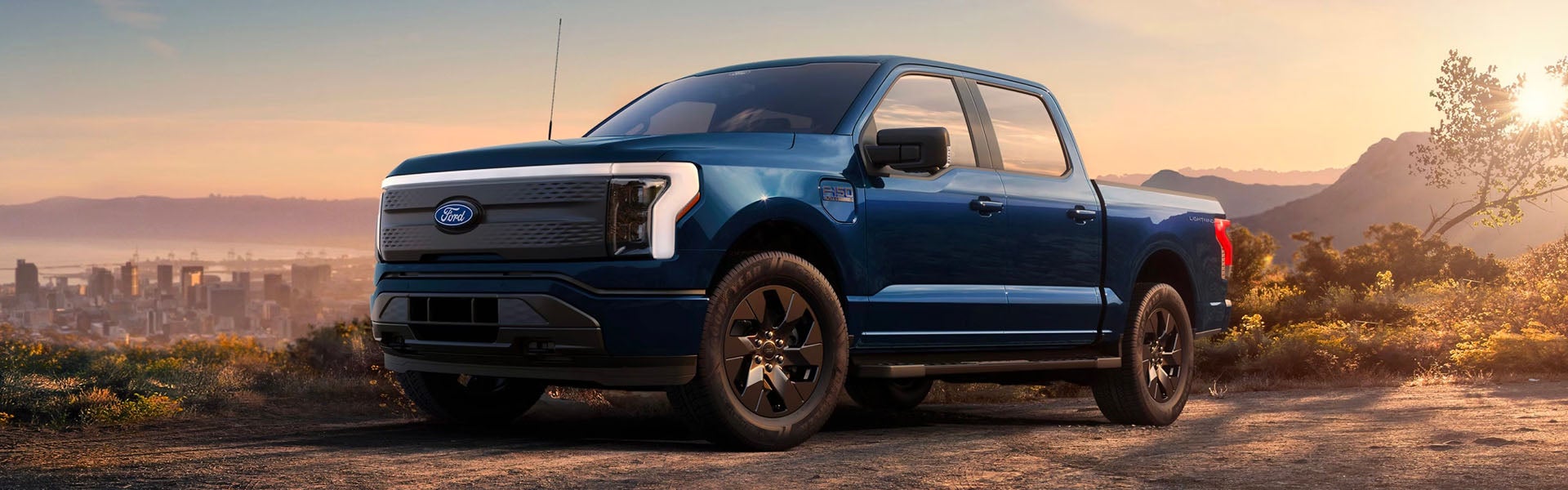Blue Ford F-150 Lightning electric truck parked on a hill overlooking a city and mountains at sunset.
