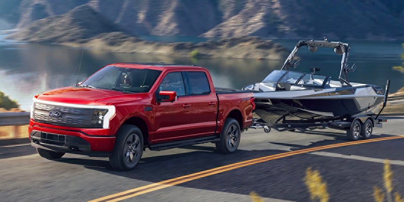 Red Ford F-150 Lightning electric truck towing a boat on a trailer along a scenic road beside a lake and mountains.