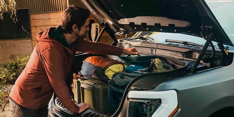 Man reaching into the open front trunk (frunk) of a vehicle, which is packed with bags and gear.