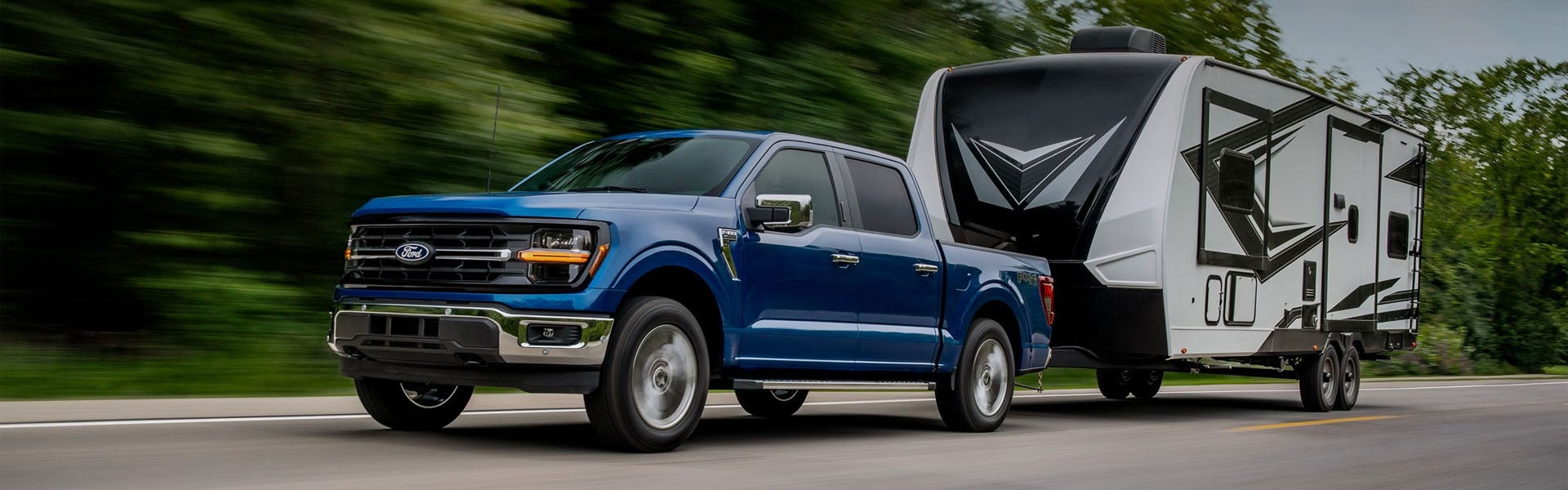 Blue Ford F-150 pickup truck towing a white and black travel trailer on a road, blurred trees in background.
