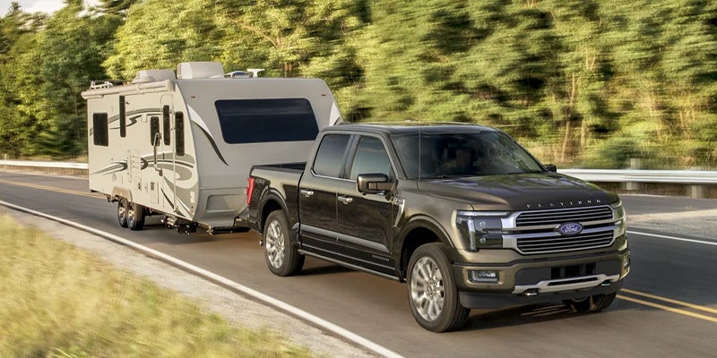 Dark gray Ford F-150 Platinum pickup truck towing a white travel trailer on a highway with blurred green trees