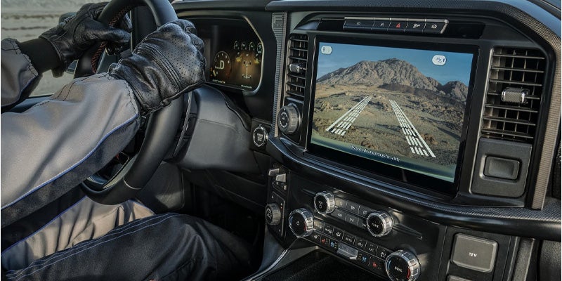 Gloved hands on a steering wheel, with a large infotainment screen showing a rear camera view of rocky terrain and guiding lines