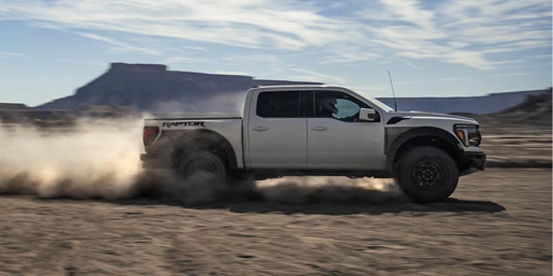 White Ford Raptor pickup truck driving off-road, kicking up a large cloud of dust in a desert landscape.