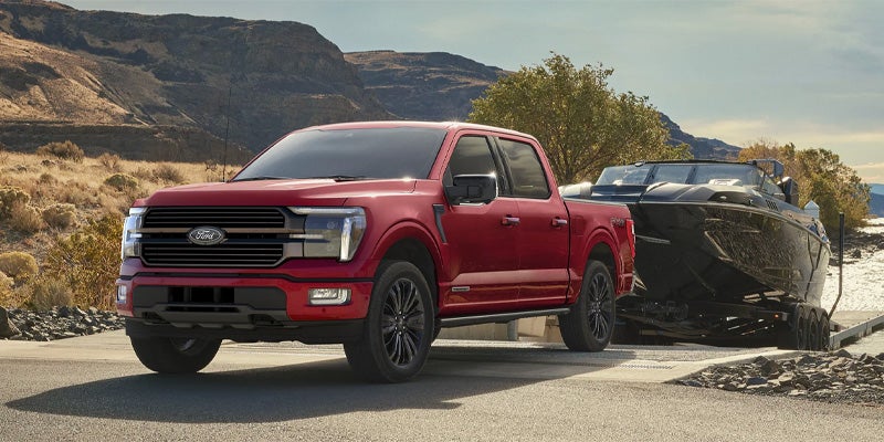 Red Ford F-150 truck parked on a boat ramp, towing a black boat on a trailer, with mountains in the background