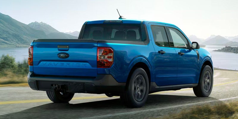 Blue Ford Maverick pickup truck driving on a scenic road with mountains and a lake in the background