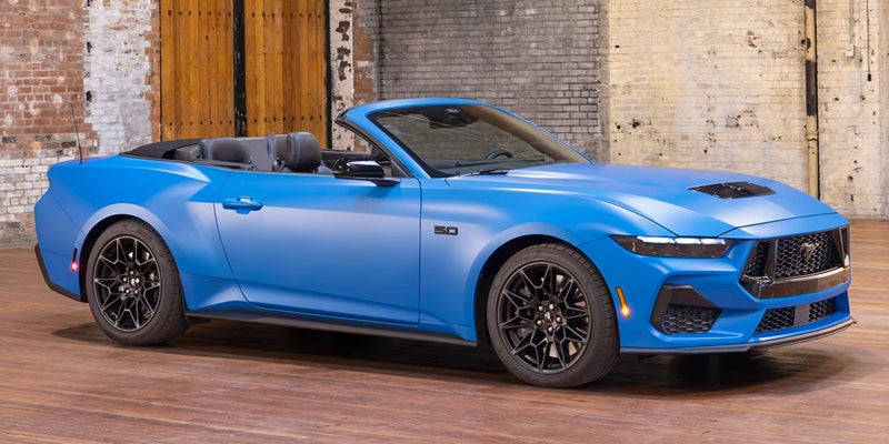 Blue Ford Mustang convertible with its top down, parked indoors with brick walls and wooden floors in the background.