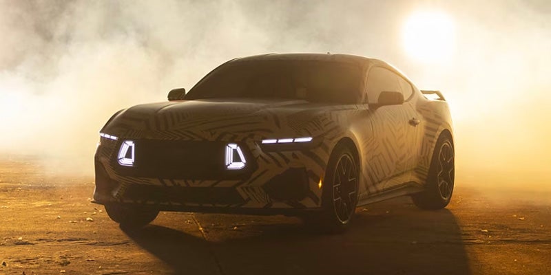 Camouflaged prototype sports car with distinctive LED headlights, partially obscured by smoke and sunlight.