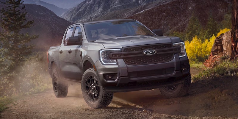 Gray Ford pickup truck driving on a rugged mountain trail, kicking up dust with forested hills and rocky peaks in the background