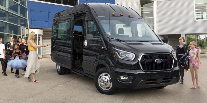 Black Ford Transit passenger van with side door open, an adult and group of children approaching/leaving the van.