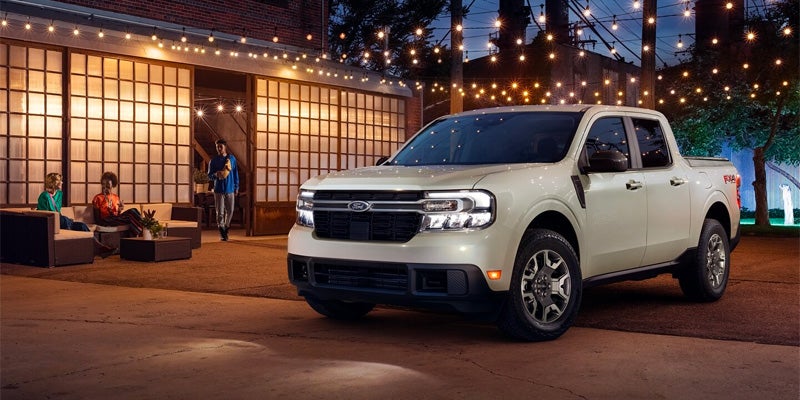 Cream-colored Ford Maverick pickup truck parked at night in an outdoor urban setting with people relaxing and string lights.