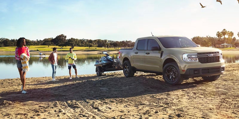 Tan pickup truck towing a jet ski on a sandy lake shore with three people and birds flying overhead.