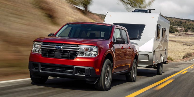 Red Ford Maverick pickup truck towing a small silver travel trailer on a winding road with blurred background.