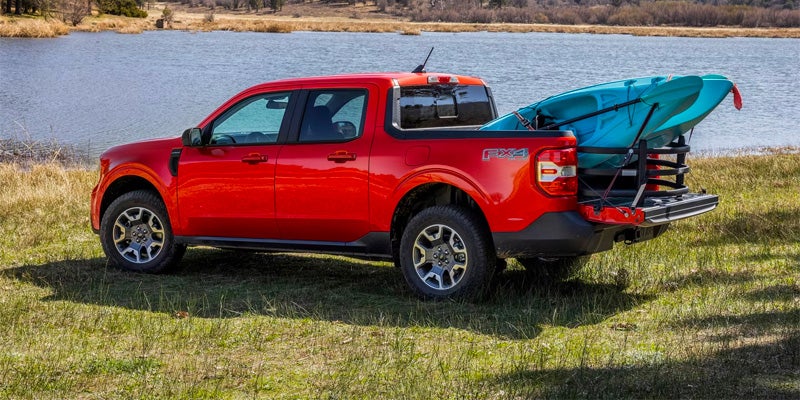 Red pickup truck with a kayak in its bed, parked on grassy ground beside a lake.