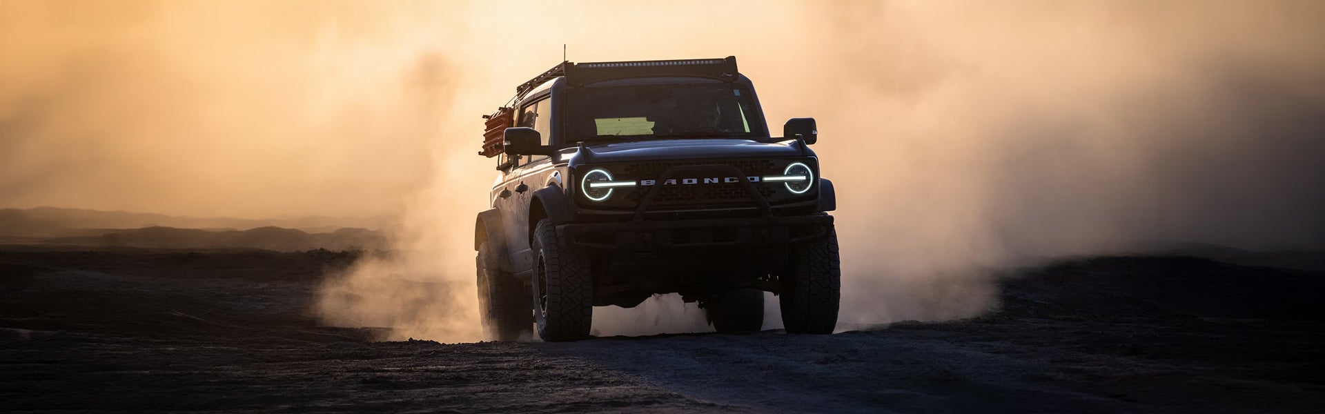 Dark Ford Bronco SUV with glowing headlights drives on a dusty road at sunset, kicking up a cloud of dust.