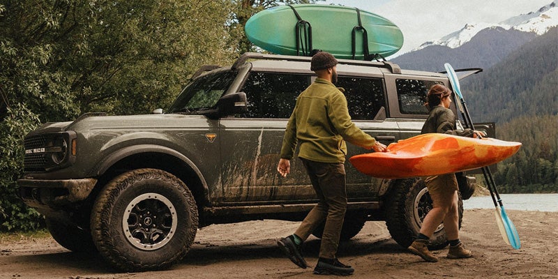 Muddy green Bronco with surfboard on roof, couple carrying kayak by a lake and mountains.