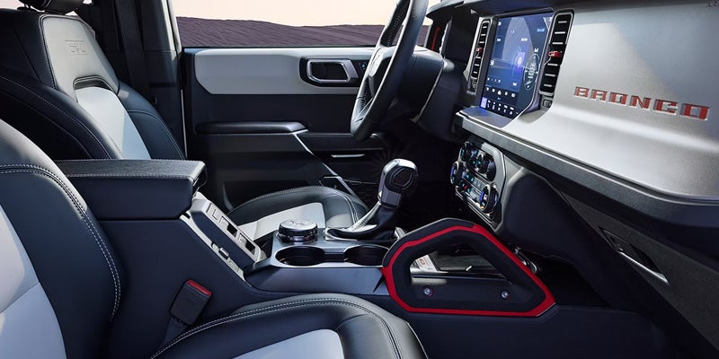 Interior of a Ford Bronco, showing black and white leather seats, center console, dashboard with screen, and red grab handle.