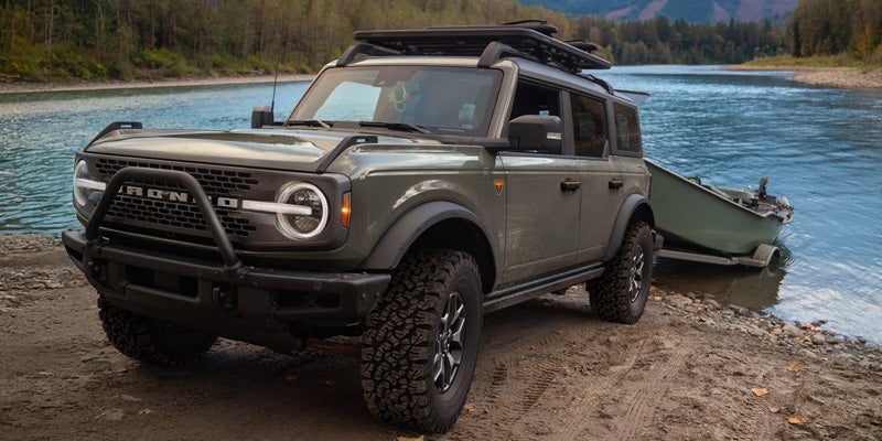 Green Ford Bronco SUV with a roof rack and brush guard, parked on a muddy riverbank next to a boat trailer partially in water