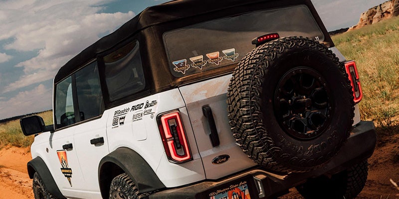 Rear view of a white Ford Bronco SUV with a soft top and spare tire on a sandy off-road trail