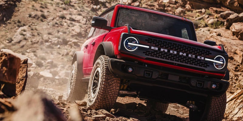 Red Ford Bronco SUV driving on a rocky, dusty off-road trail, angled towards the viewer
