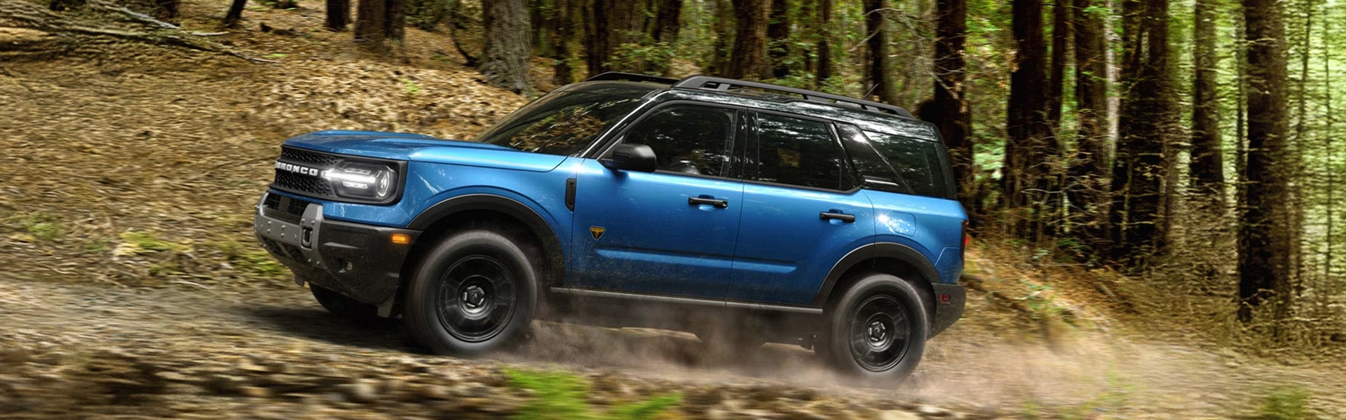 Blue Ford Bronco Sport SUV kicking up dust as it drives on a dirt path through a sun-dappled forest.