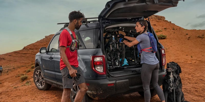 Man and woman loading bikes into an SUV's trunk, with a black dog nearby, in a desert landscape.