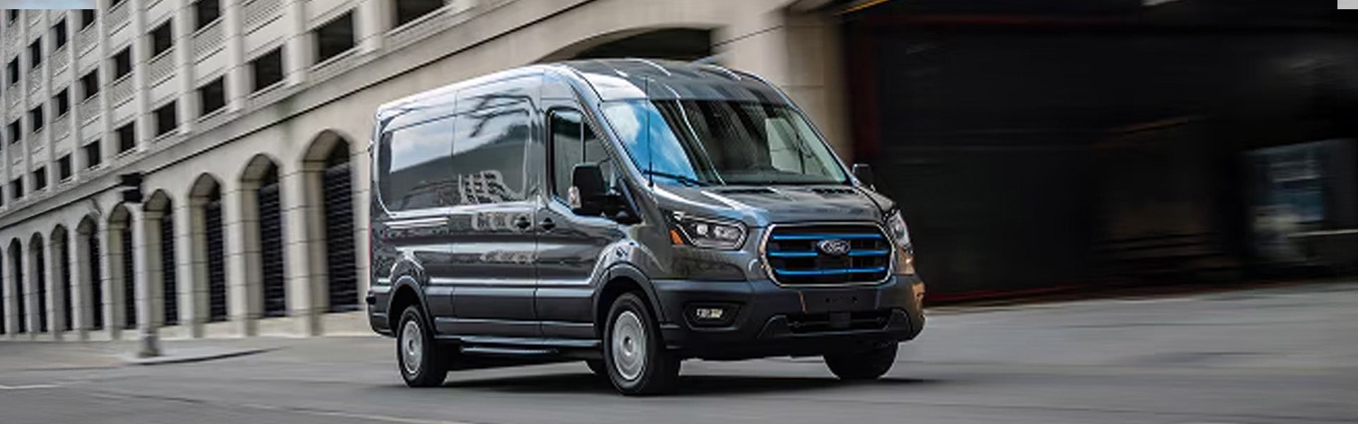 Dark gray Ford E-Transit cargo van driving on a city street with building arches in the background.