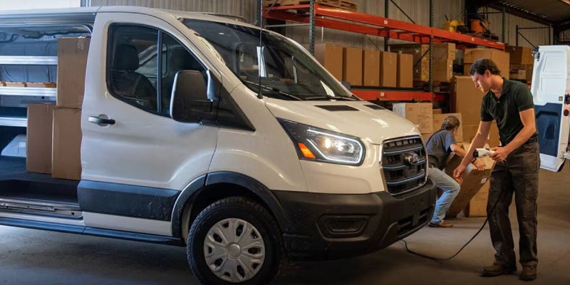 White electric delivery van being charged by a man and loaded with boxes by another in a warehouse.