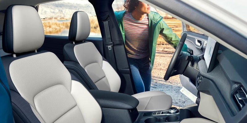 Car interior with two-tone light and dark seats, dashboard, and a person leaning into the open driver's door.