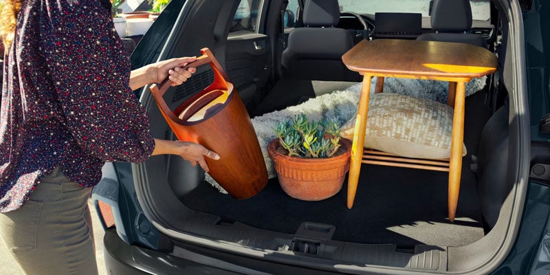 Person loading a wooden carrier into a car trunk already containing a small table, potted plant, and blanket.