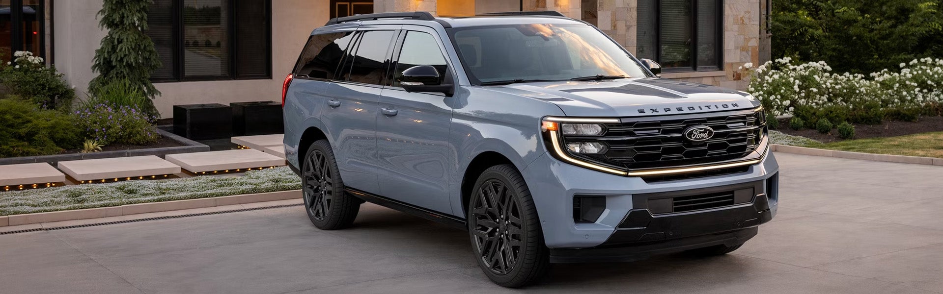 Gray-blue Ford Expedition SUV with black grille and wheels parked in a modern driveway next to a house and landscaping.