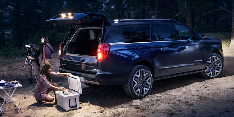 Dark SUV with open trunk lights up a family stargazing and accessing a cooler in a forest at night