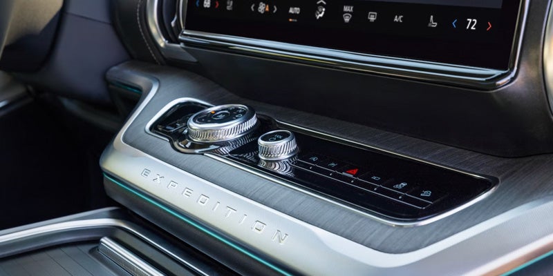 Car interior console with silver rotary dials, buttons, and EXPEDITION branding on a textured silver panel.