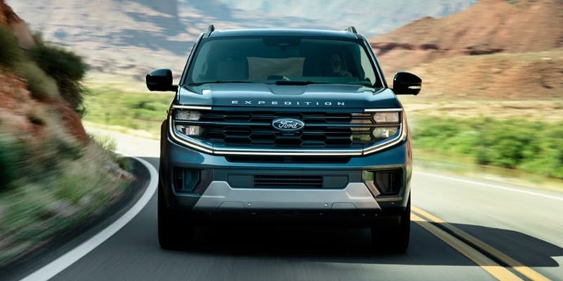 Front view of a dark blue Ford Expedition SUV driving on a winding road through a mountainous, desert-like landscape.