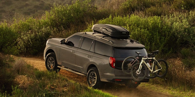 Dark gray SUV with roof cargo box and two bicycles on a dirt path, surrounded by green foliage.