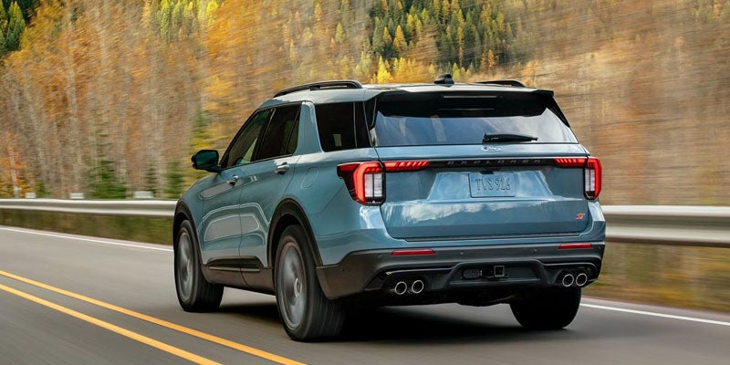 Teal Ford Explorer SUV driving on a road with autumn trees in the background, shown from a rear-quarter view.