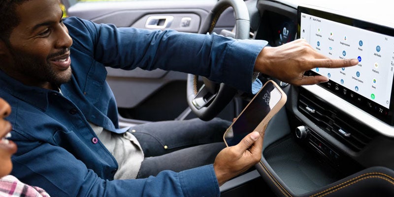 A man smiles while holding a phone and touching a large in-car infotainment display with apps