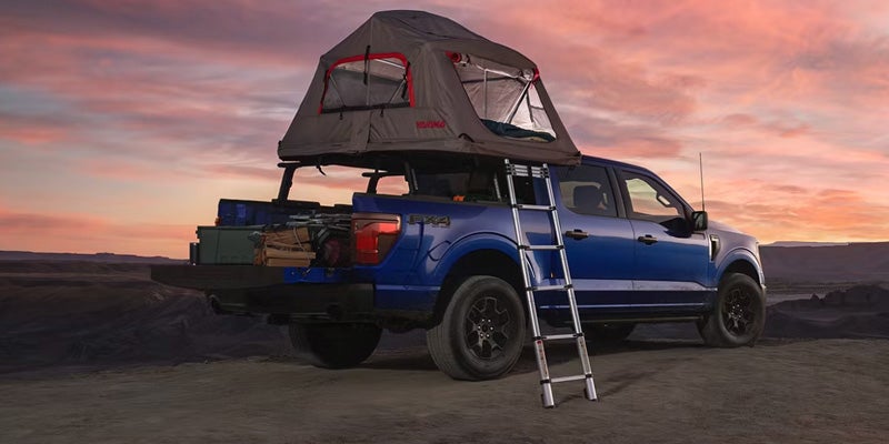 Blue pickup truck with a rooftop tent and ladder, camping gear in its bed, parked in a desert at sunset
