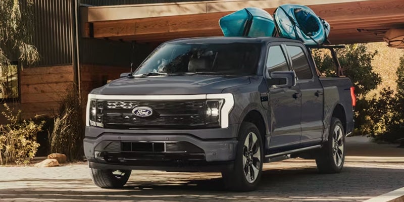 Gray Ford F-150 Lightning electric pickup truck with two blue kayaks on a roof rack, parked next to a modern building.