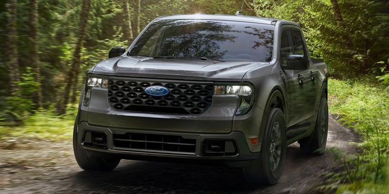 Dark gray Ford Maverick pickup truck driving on a dirt road through a forest